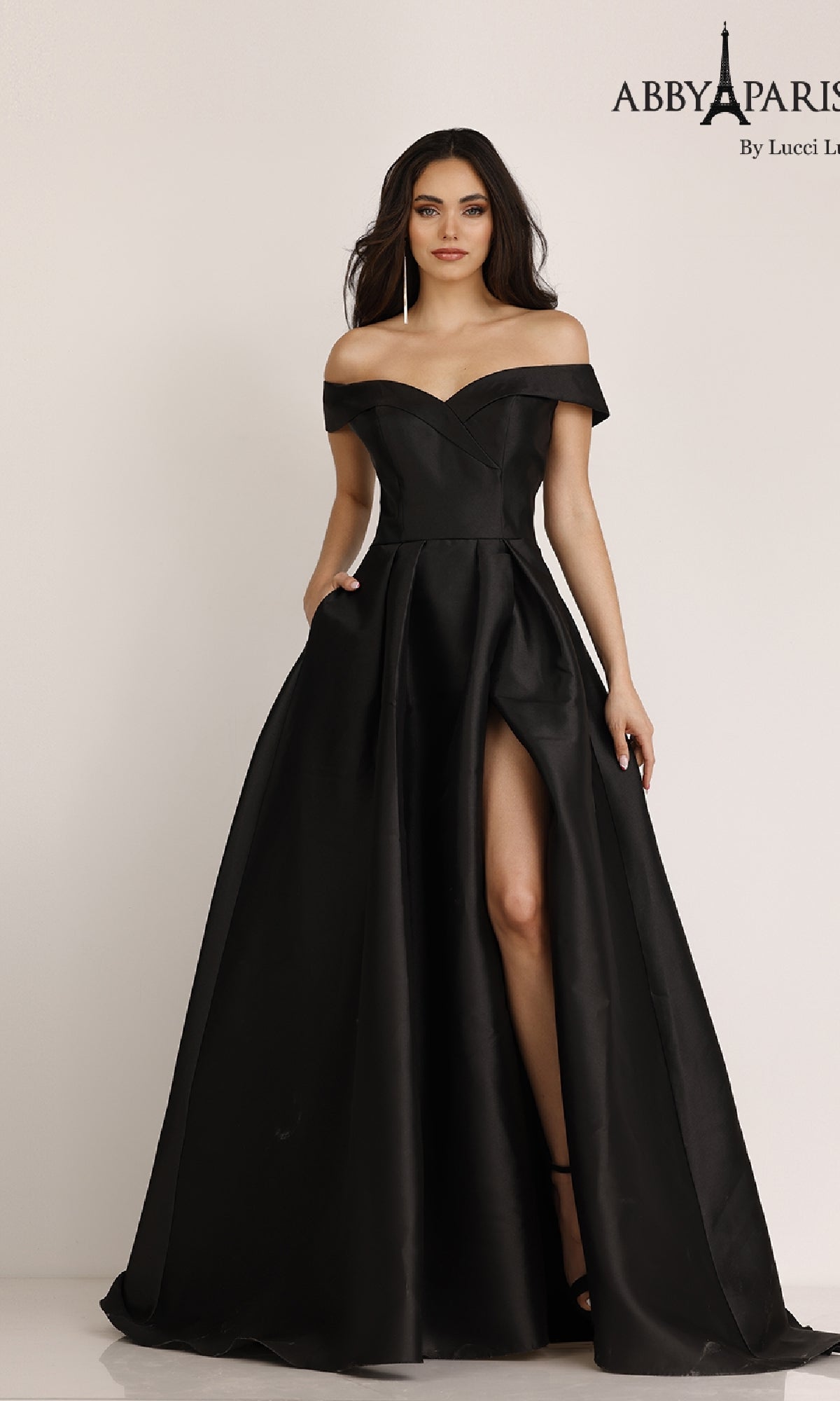 A woman stands against a plain background wearing the Classic Mikado Prom Ball Gown Abby Paris 90023—an off-the-shoulder, floor-length black dress with a thigh-high slit.